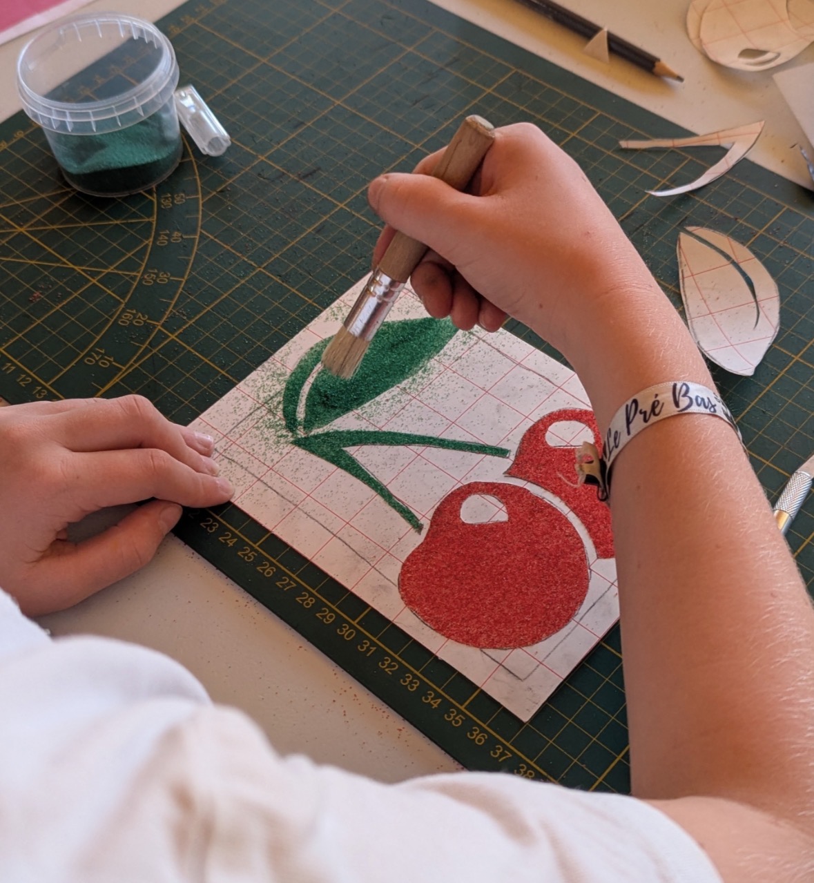 Gros plan sur la main d'un enfant appliquant au pinceau du sable coloré vert sur les feuilles d'un tableau représentant des cerises.