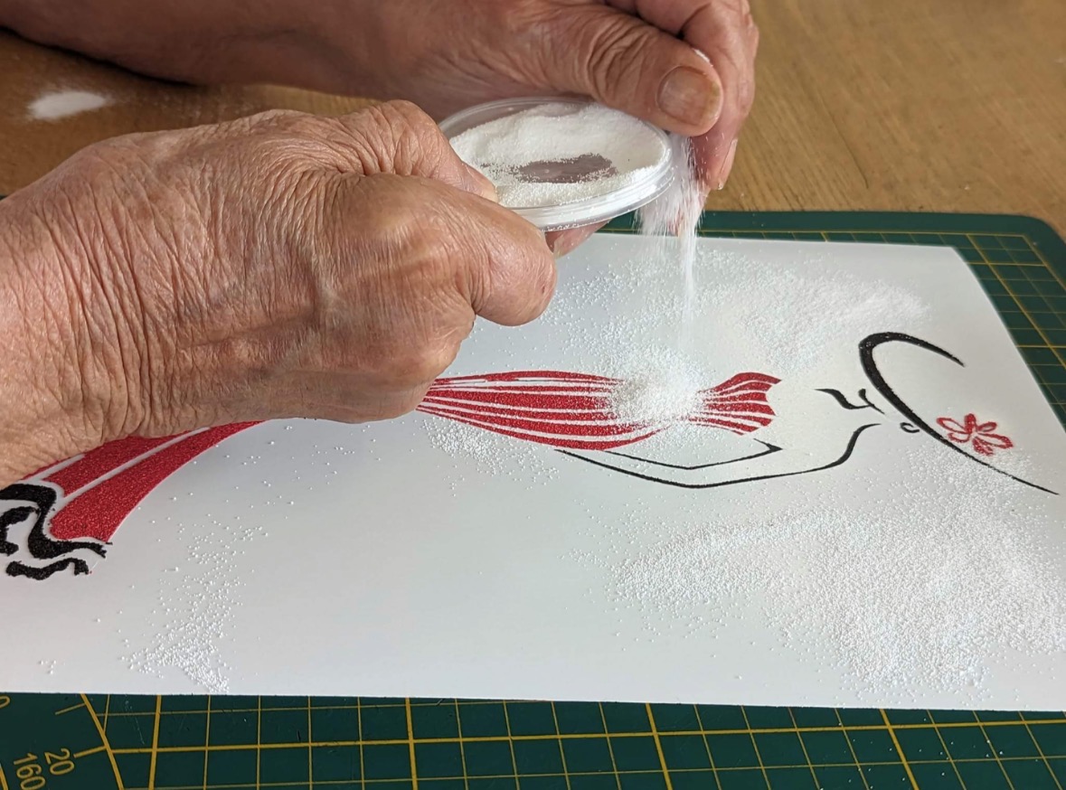 Gros plan sur des mains versant du sable blanc pailleté sur le contour dessiné d'une sirène stylisée, dont la queue est déjà recouverte de sable rouge.