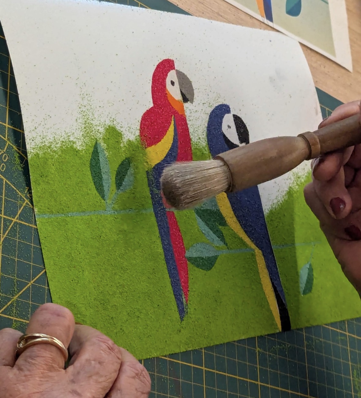 Gros plan sur des mains appliquant du sable coloré au pinceau sur un tableau représentant deux aras rouge et bleu sur fond vert, posé sur un tapis de découpe.